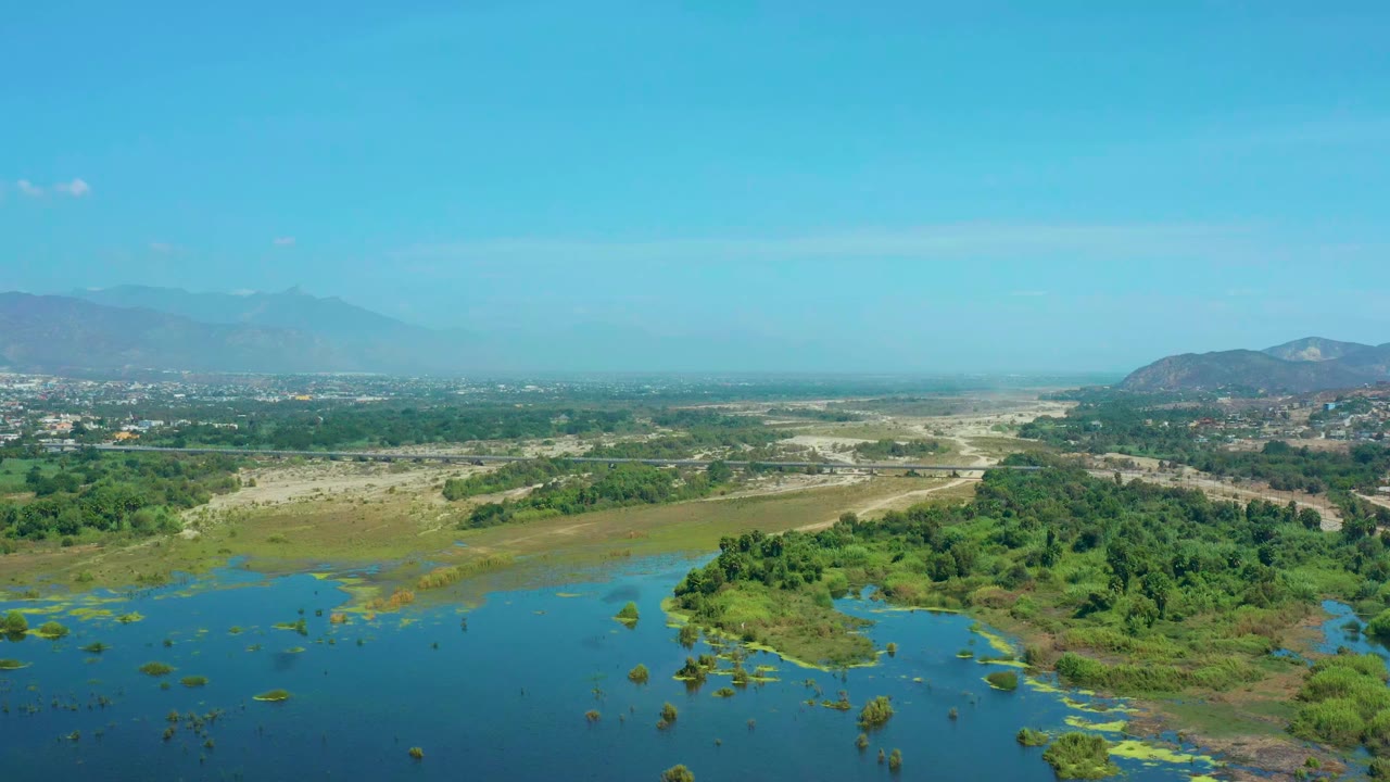 Aerial view one second into the San Jose del Cabo marshland clip, with blue water in front, a green thicket to the right, pale dry ground beyond, and no visible people.
