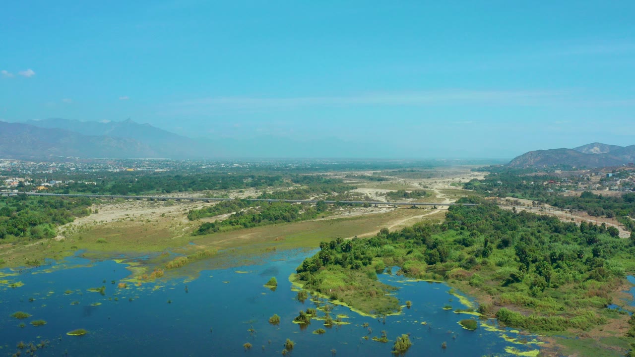 Opening aerial frame over San Jose Del Cabo, Mexico, showing blue water and green wetland vegetation below dry terrain and hazy mountains, with no visible people.