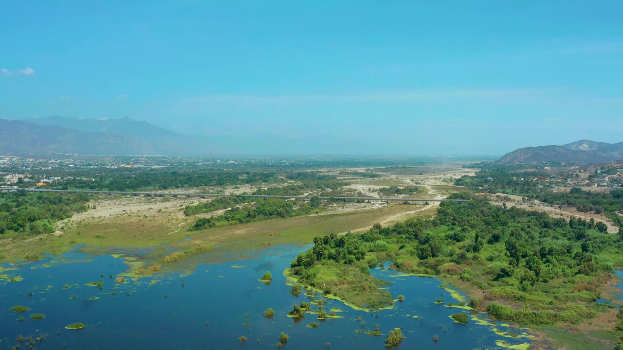Early aerial frame from San Jose Del Cabo, Mexico, with blue water and green patches in the foreground, dry flats through the center, and distant mountains under clear sky, with no visible people.