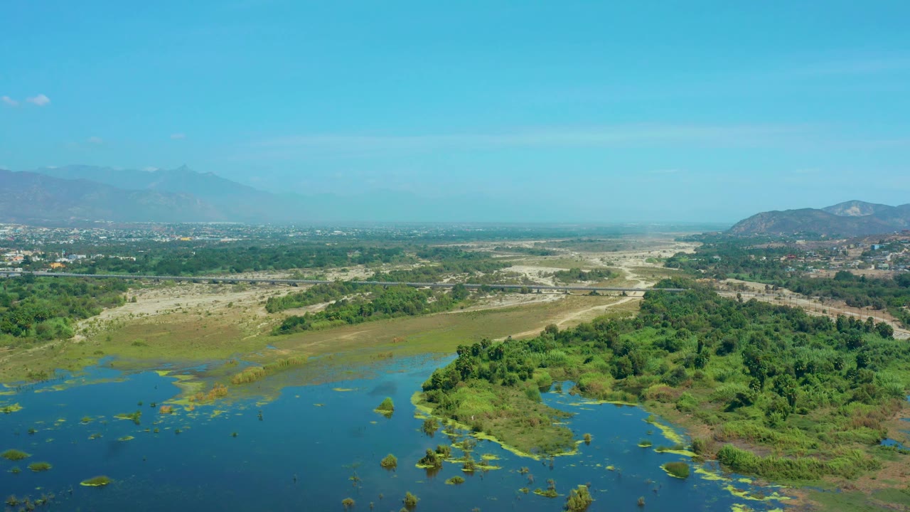 Aerial frame over San Jose Del Cabo, Mexico, with a green vegetated mound near the lower right, water across the lower left, dry terrain beyond, and hazy mountains in back, with no visible people.