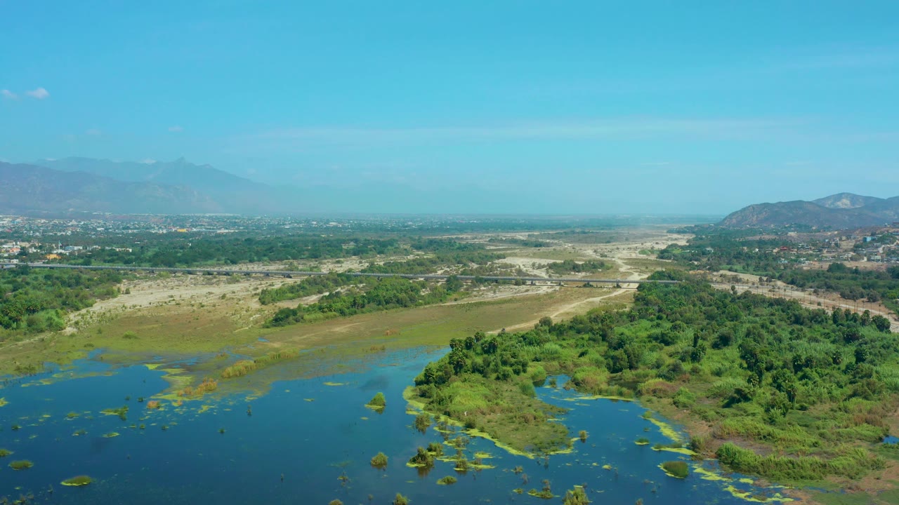 Mid-clip aerial view in San Jose Del Cabo, Mexico, looking across blue wetland water and green vegetation toward tan flats, scattered roads, and a soft mountain horizon, with no visible people.