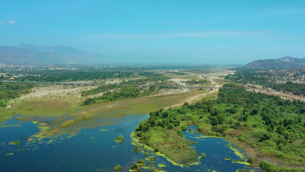 High aerial frame over San Jose Del Cabo showing wetland water edged by green brush, tan open ground, faint roads, scattered distant buildings, and no visible people.