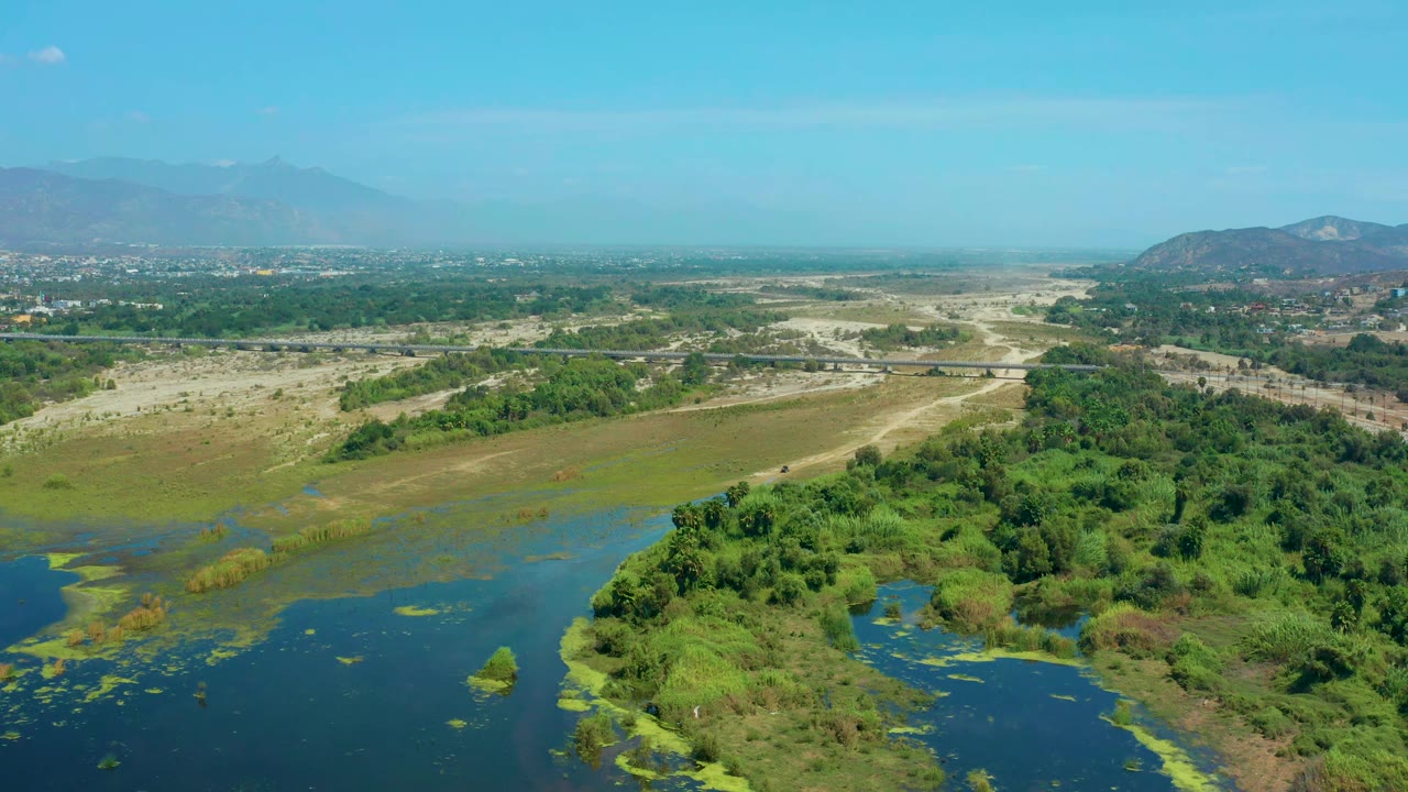 Aerial view in San Jose Del Cabo, Mexico, with a darker water channel at lower left, dense green brush through the center, pale sandy ground beyond it, distant hills, and no visible people.