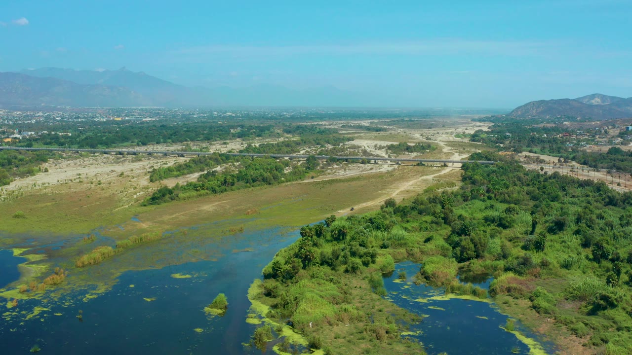 Wide aerial frame over San Jose Del Cabo, Mexico, showing open water at lower left, green vegetation along the lower right, sandy terrain across the middle, distant settlement on the horizon, and no visible people.