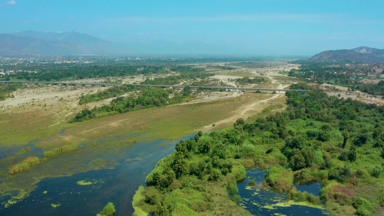 Opening frame of a wide aerial view over green wetland vegetation and dark water near San Jose Del Cabo, Mexico, with pale dry flats, distant hills, and no visible people.