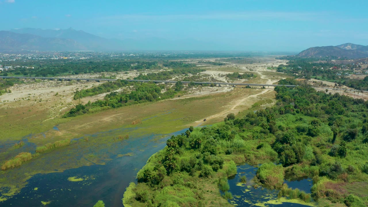 Aerial frame over wetland plants and shallow water near San Jose Del Cabo, Mexico, with a road or bridge crossing the midground and hazy mountains in the distance, no visible people.