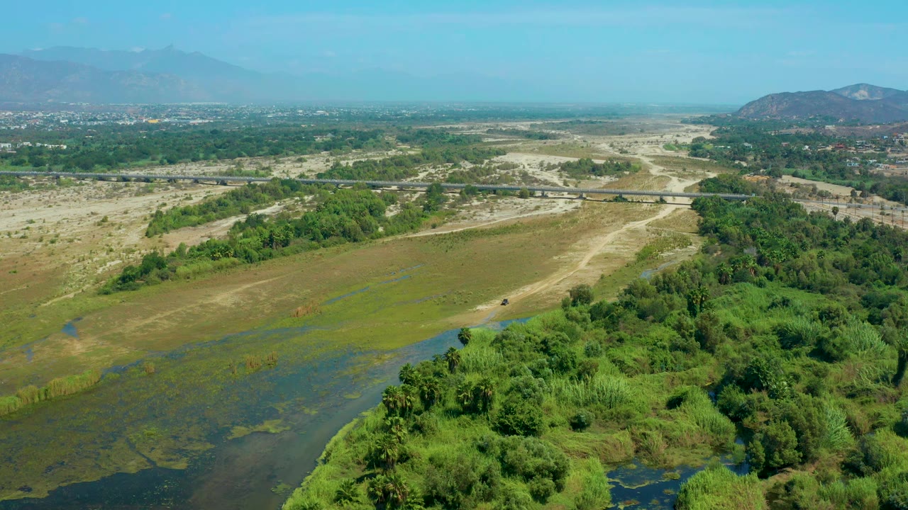 Aerial view in San Jose Del Cabo with dense green brush in the lower frame, muddy open ground and a pale road through the middle, a bridge line, distant settlement, and no visible people.