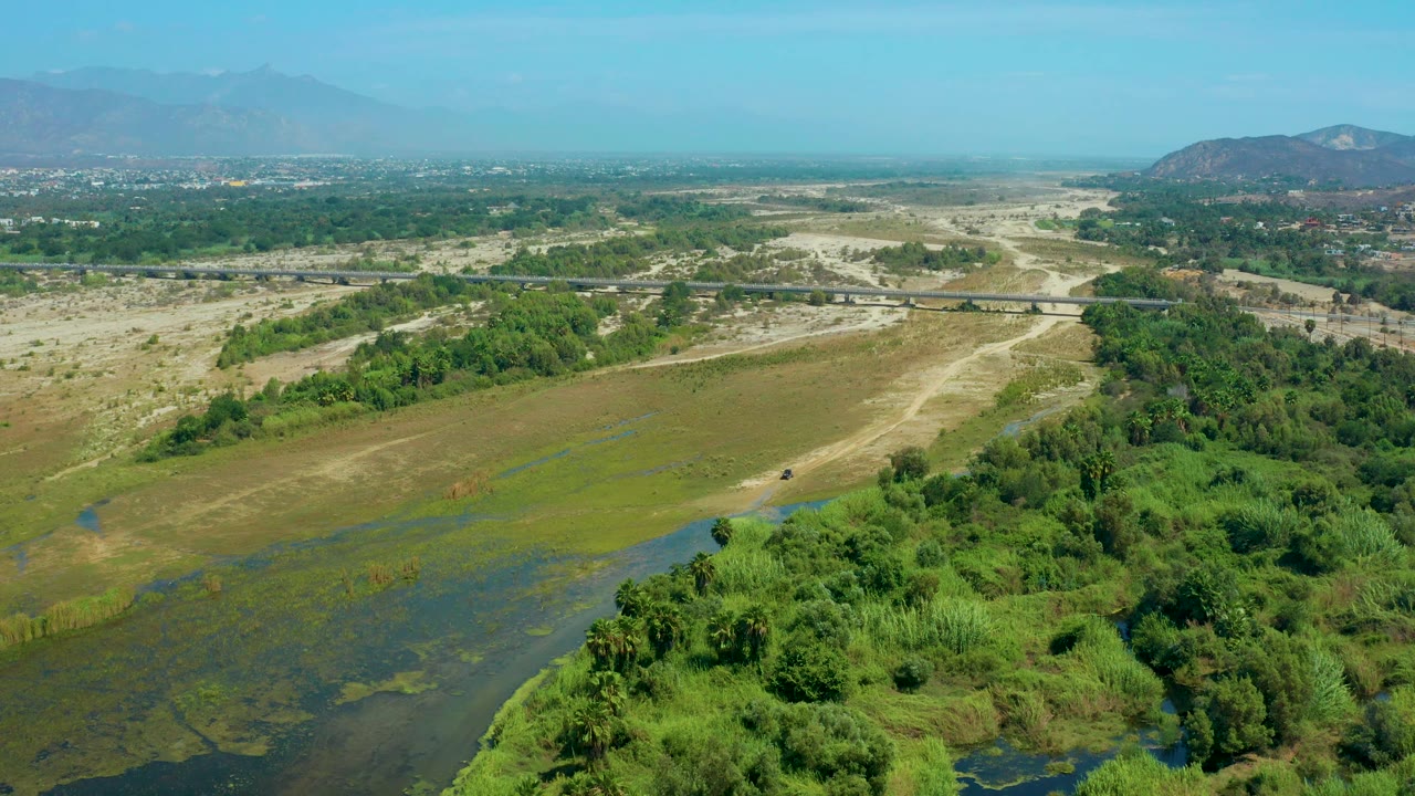 San Jose Del Cabo aerial frame showing a larger patch of green wetland vegetation at lower right, muddy track lines across open ground, a long bridge in the midground, distant buildings, and no visible people.