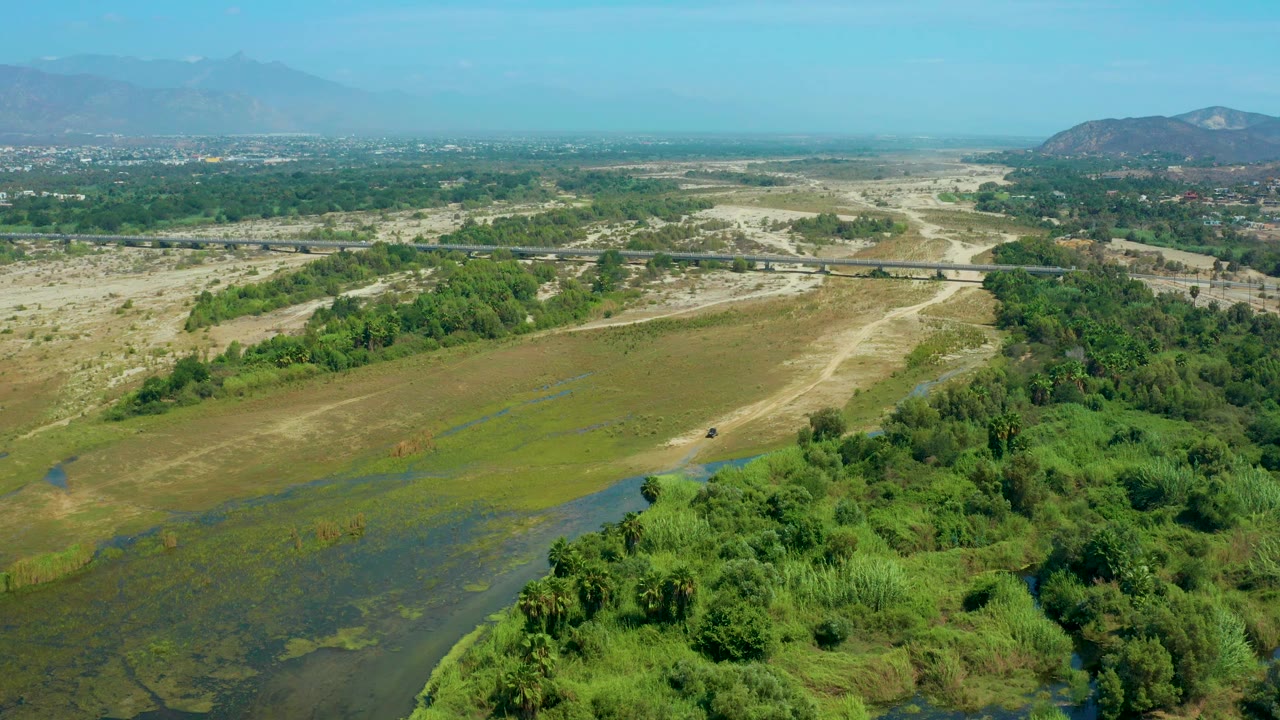 Mid-clip aerial view over San Jose Del Cabo with darker muddy water in the foreground, green trees along the edge, pale open flats, a straight bridge, haze on the mountain horizon, and no visible people.