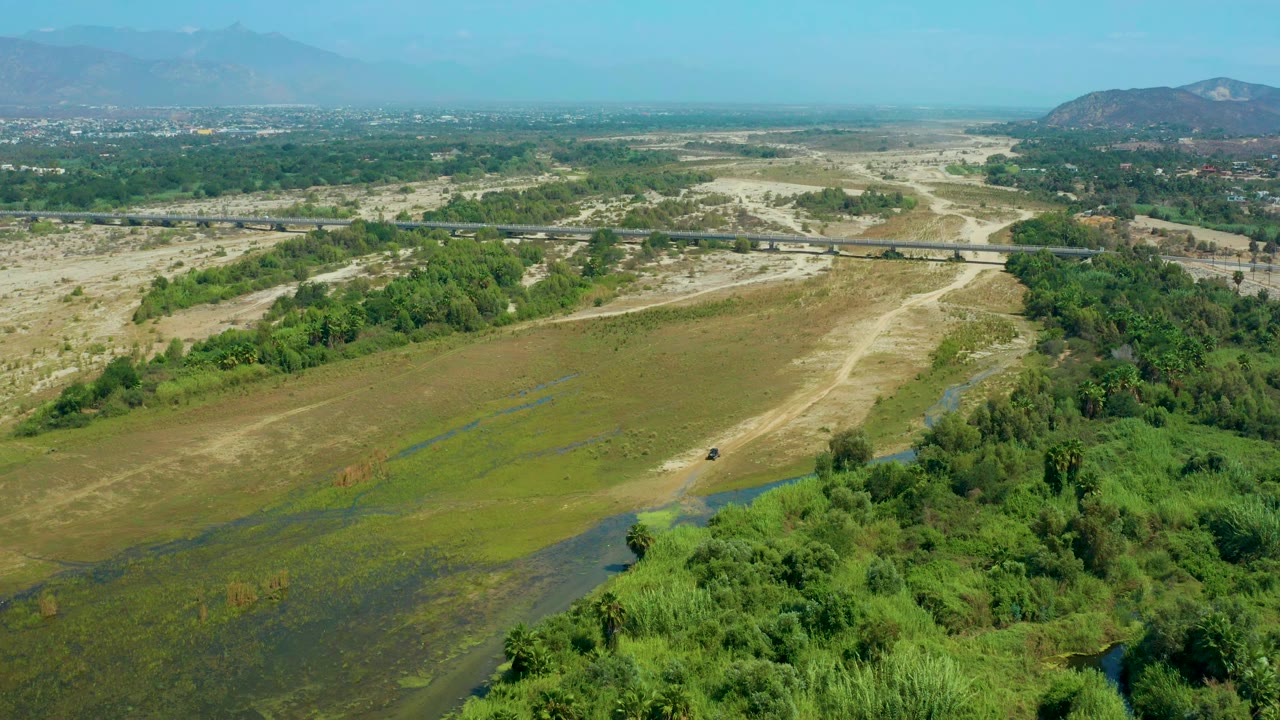 Second aerial frame from San Jose Del Cabo, Mexico, with the concrete bridge near the upper midground, muddy road lines through tan ground, dense green trees at right, and no visible people.