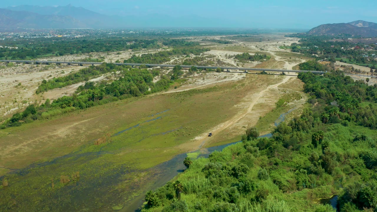 Third frame shows the San Jose Del Cabo inland terrain from above, with a band of trees along the right side, pale muddy tracks across the center, a bridge beyond them, and no visible people.
