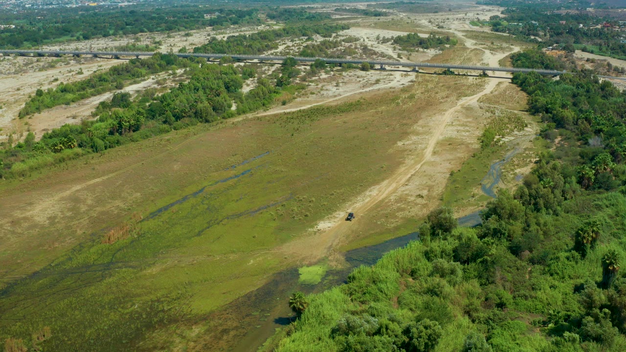 Aerial frame over San Jose del Cabo, Mexico, looking across a marshy green plain with pale sandy tracks, right-side brush, distant roads, and no visible people.
