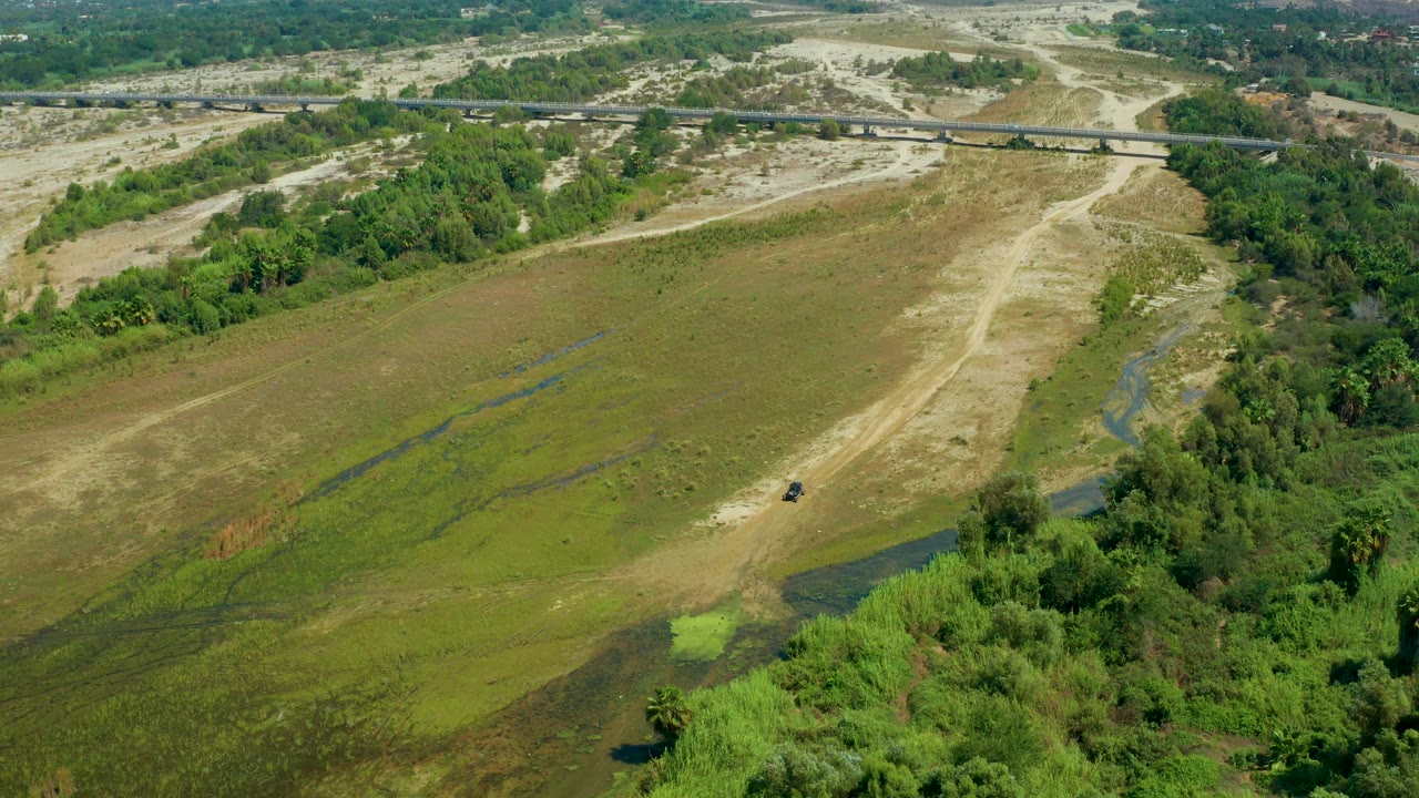 Aerial frame over San Jose del Cabo, Mexico, with a wide green field, a pale dry wash cutting upward through frame, dense trees on the right, and no visible people.