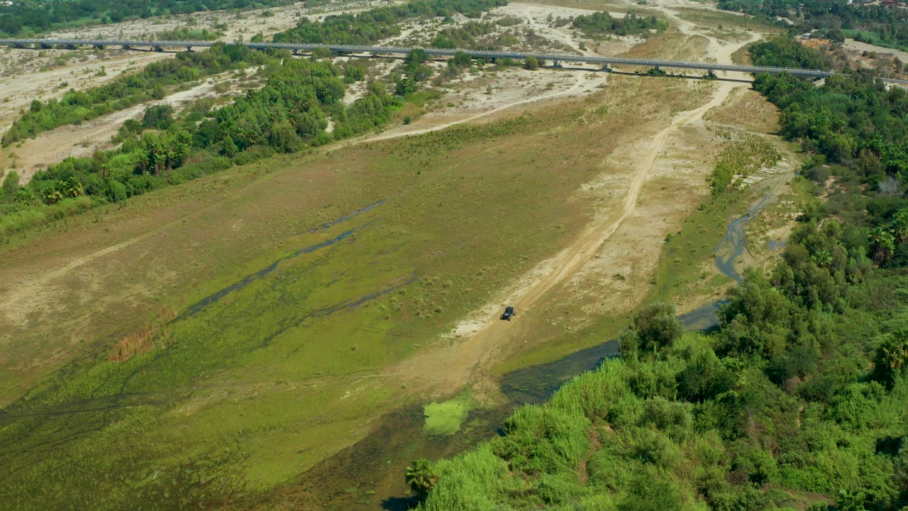 Mid-clip aerial frame over San Jose del Cabo, Mexico, showing wet green terrain, sandy channels, thick vegetation at the lower right, a road bridge beyond, and no visible people.