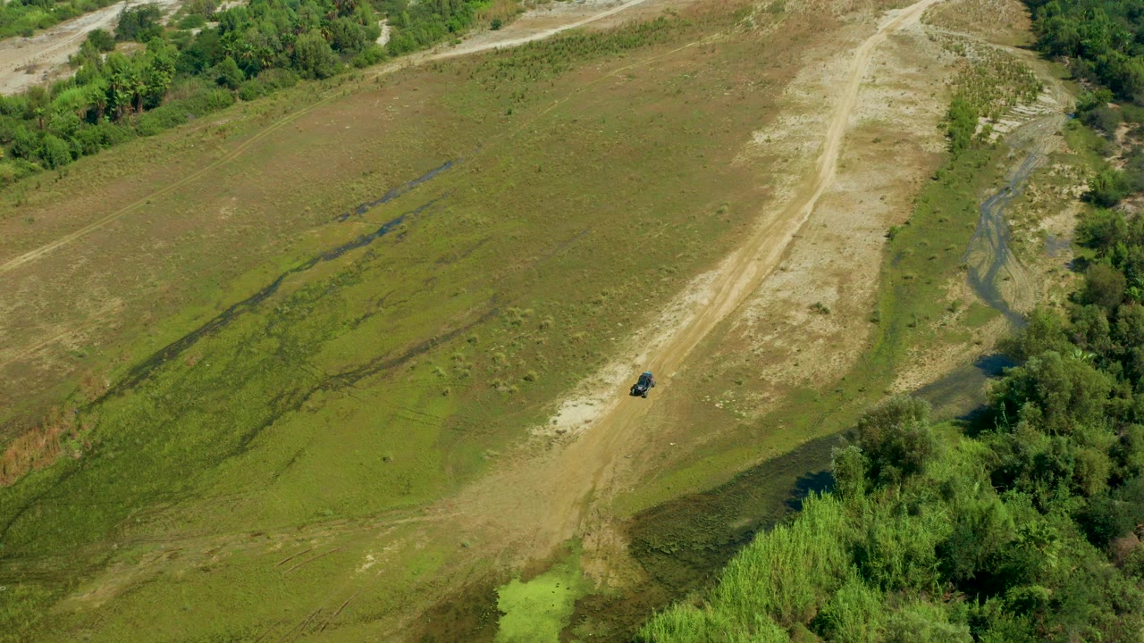 Aerial frame from San Jose Del Cabo, Mexico showing green grassland, pale dirt road, trees along the edge, and a small black car near the road with no visible people.