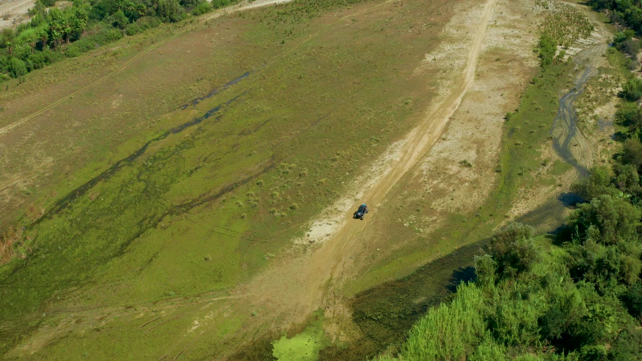 High aerial frame from San Jose Del Cabo, Mexico showing a black car below on the dirt road, a branching pale track, green low grass, and no visible people.