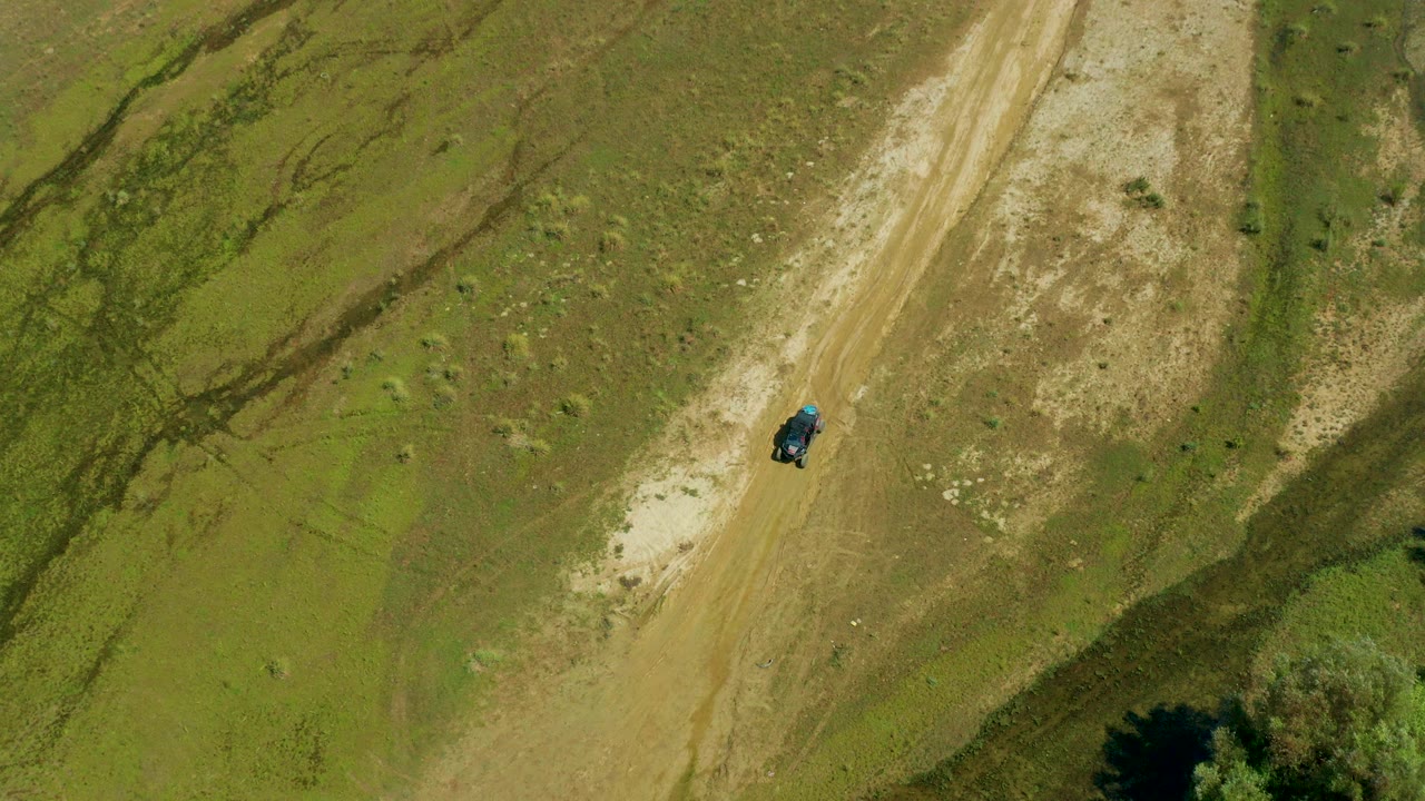 Aerial frame from San Jose Del Cabo, Mexico showing a dark vehicle on a diagonal dirt road through sparse green-brown terrain, with no visible people.