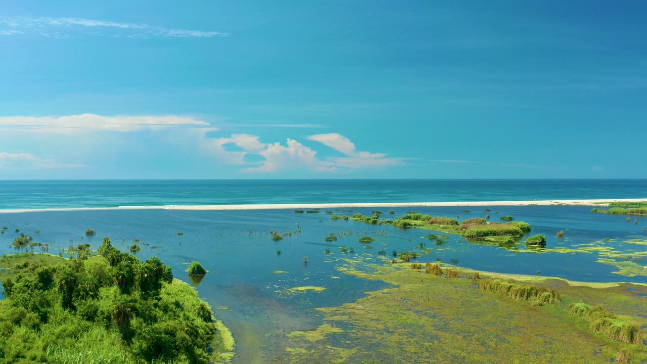 Opening frame of San Jose Del Cabo coastal water with dense green vegetation in the lower left, shallow flats through the center, blue ocean beyond, and no visible people.