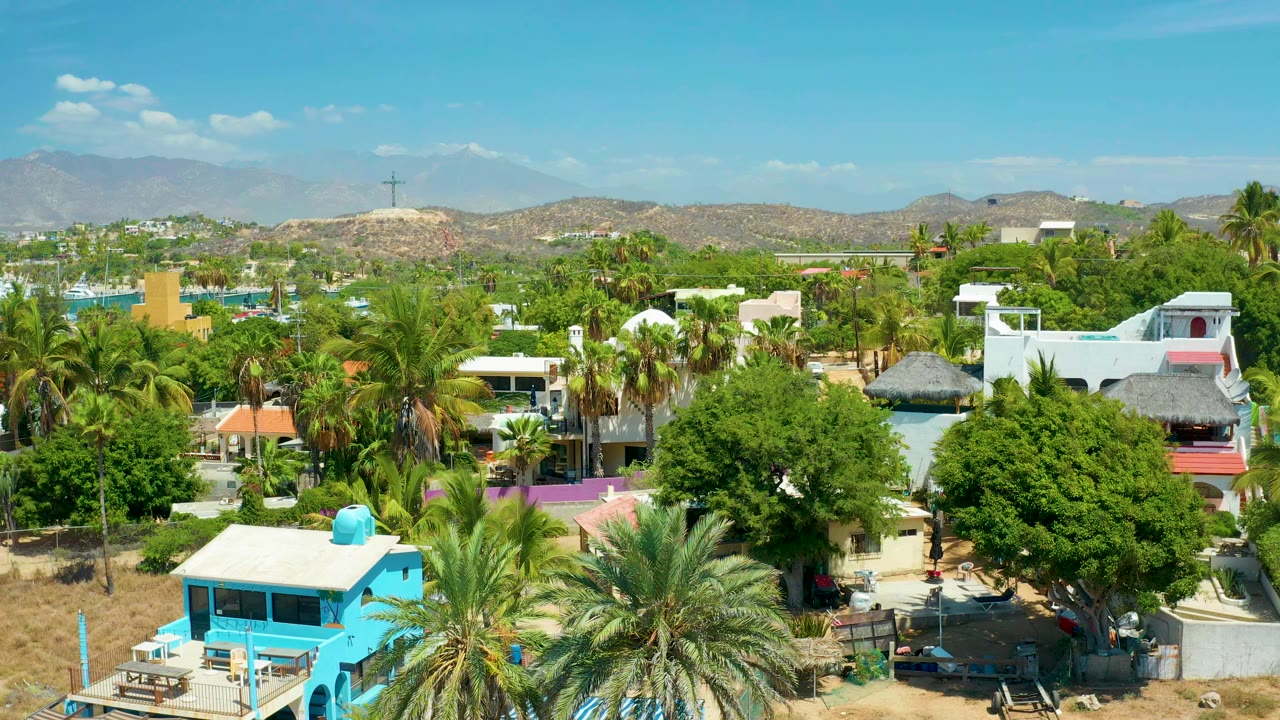 Second aerial frame in San Jose del Cabo showing white homes partly hidden by palms and large green trees, with tan hills and no visible people.