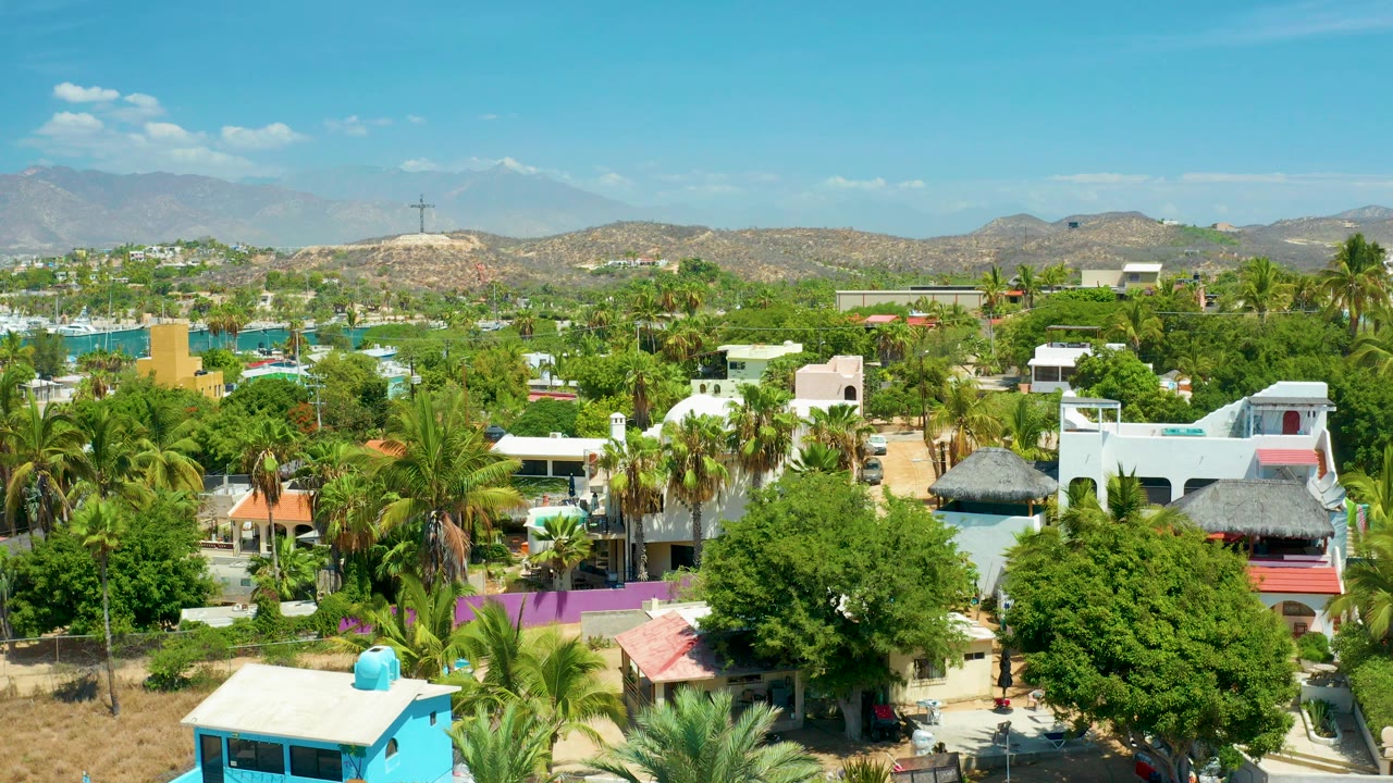 Opening aerial frame over San Jose Del Cabo, Mexico, with palm trees, pastel rooftops, turquoise water at left, hazy hills in the background, and no visible people.