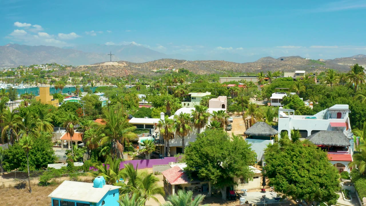 Aerial frame over San Jose Del Cabo showing white houses, a red roof, thatched roof structures, palms, turquoise water to the left, mountains beyond, and no visible people.