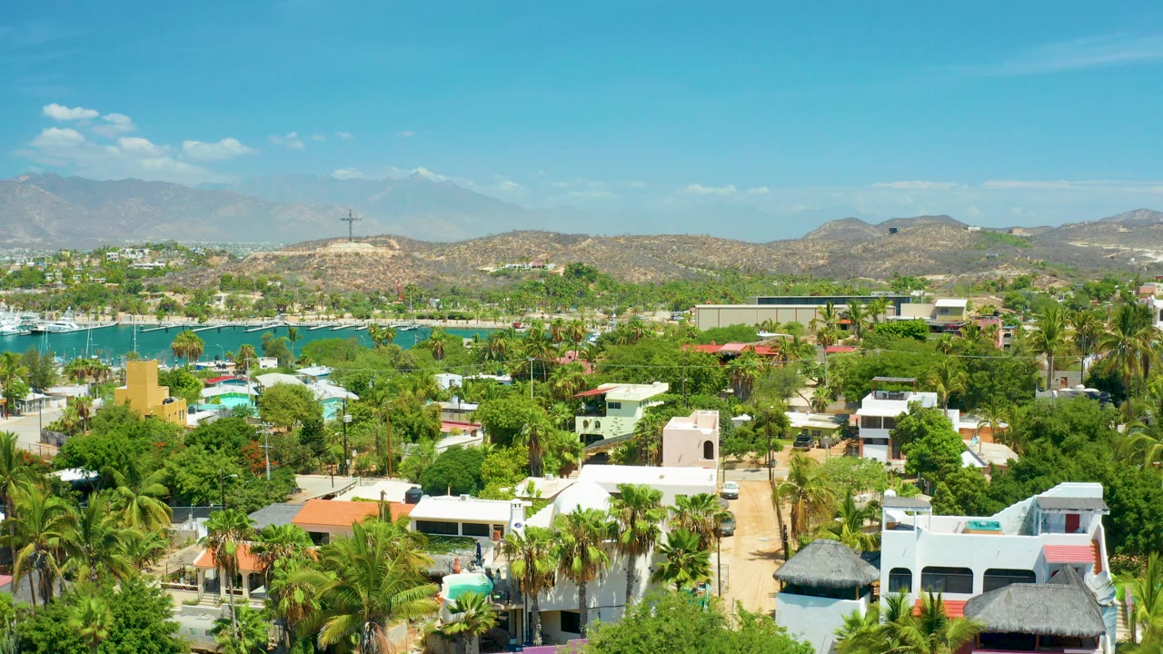 Aerial view over San Jose Del Cabo villas and palm trees with turquoise water, boats, dry hills, and mountains in the distance, with no visible people.