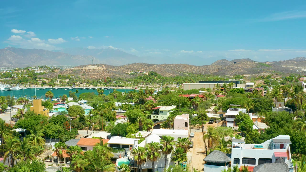 Aerial view in San Jose Del Cabo showing a palm-lined villa area, bright turquoise water with boats, low dry hills, and a clear blue sky, with no visible people.