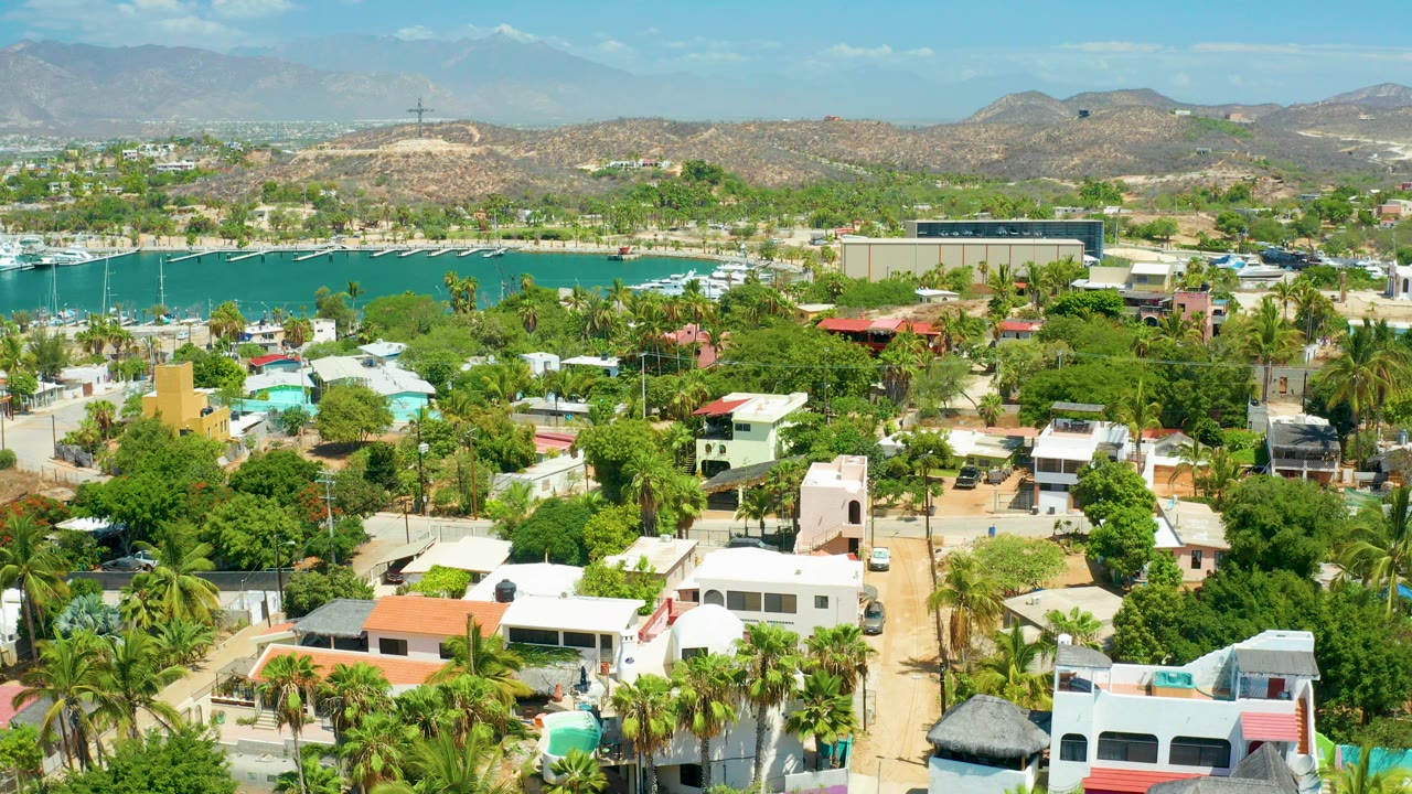 Aerial view over San Jose del Cabo with foreground homes and palms, turquoise harbor water with boats, dry hills in the distance, and no visible people.