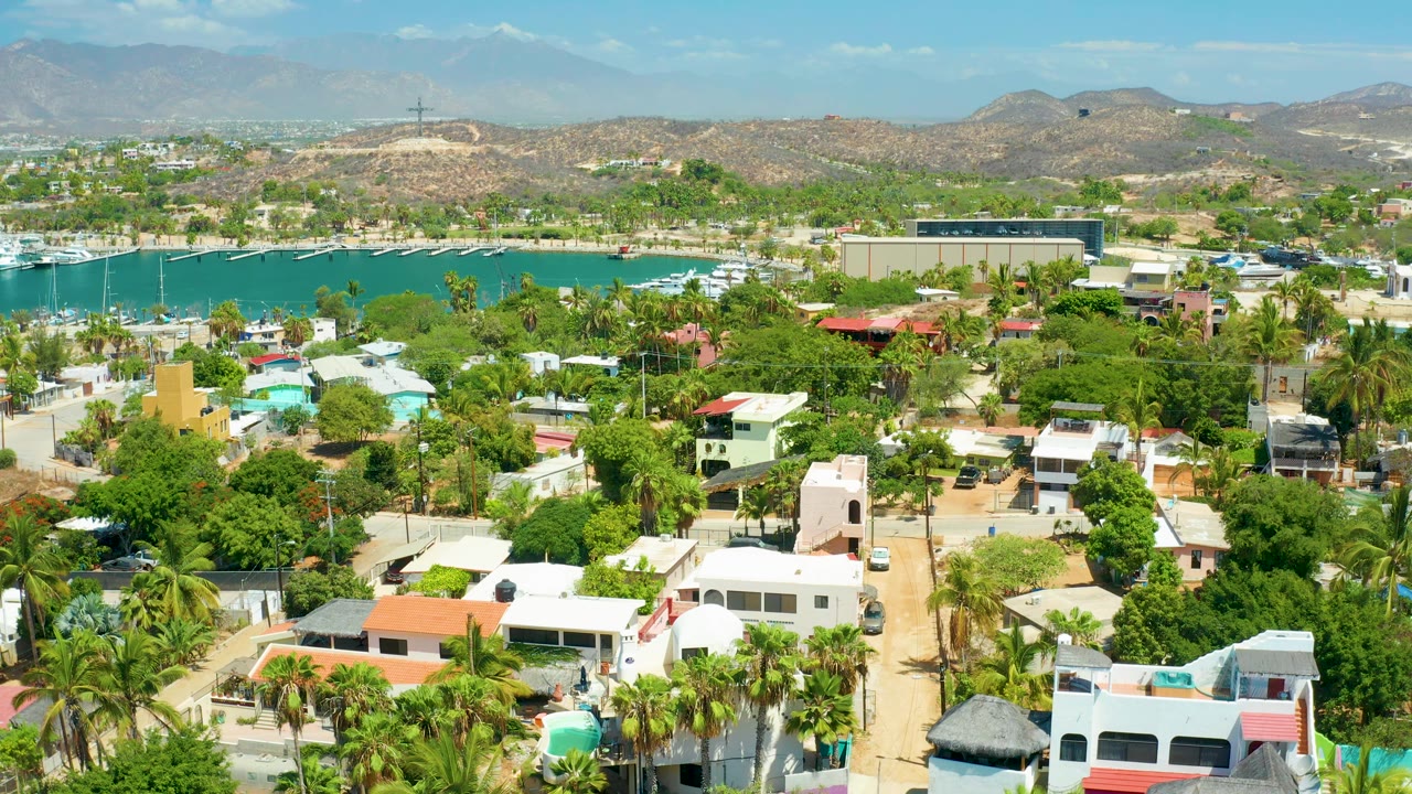 Wide drone frame in San Jose del Cabo showing white and pastel rooftops, dense green trees, marina water, small boats, distant hills, and no visible people.