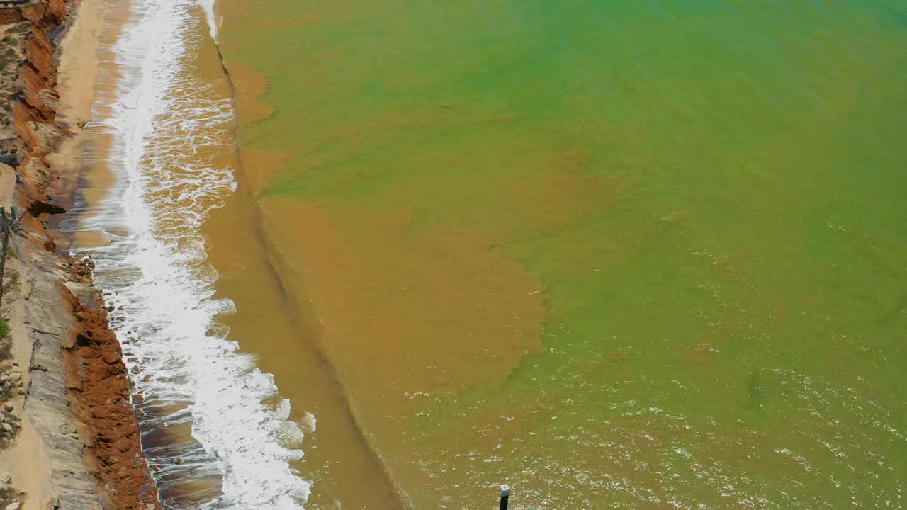 White waves run along rocky cliffs beside tan and green water.