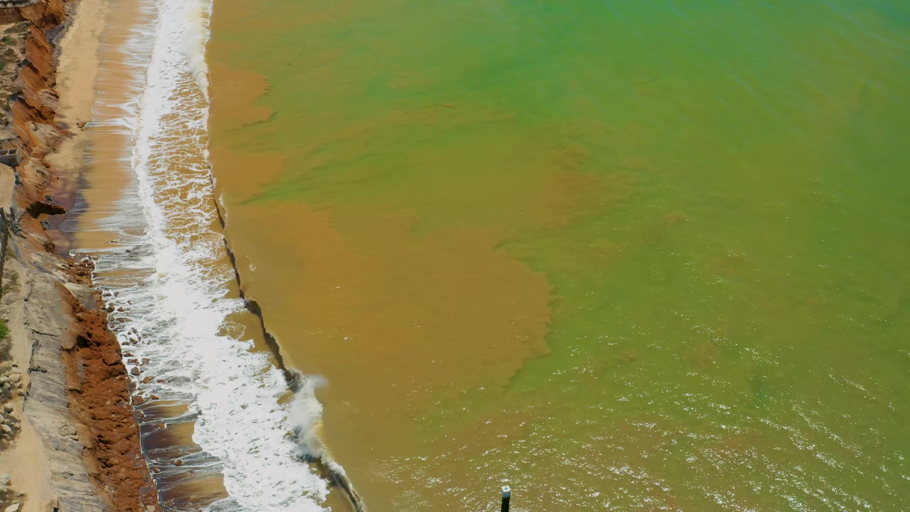 Drone-like overhead view in San Jose Del Cabo, Mexico with surf curling along the left cliff edge and a large tan sandy patch spreading through green water, with no visible people.