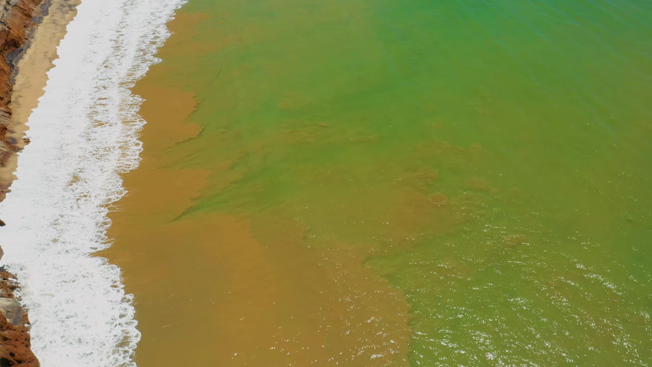 White waves run down the left side of the frame while green and tan shallow water spreads across the San Jose Del Cabo beach area, with no visible people.