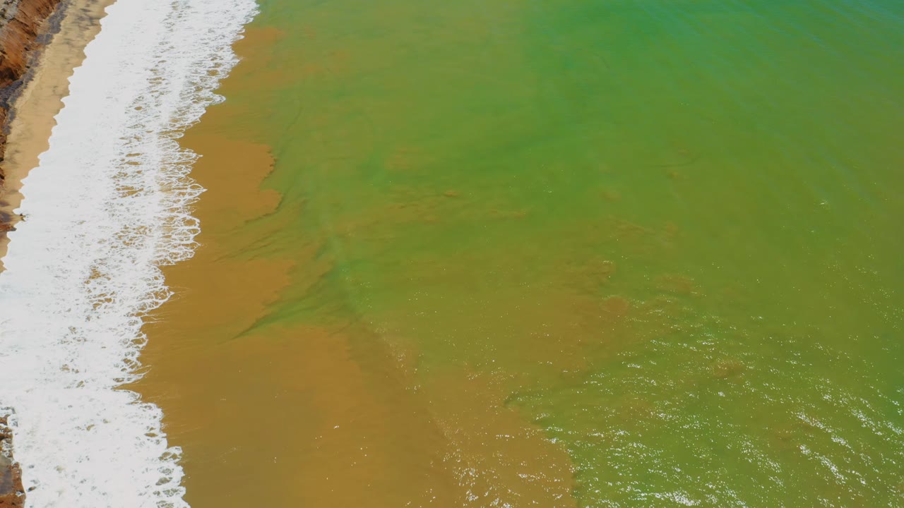 Aerial frame with a bright foam line on the left, green sea to the right, and tan sand color visible under the water near San Jose Del Cabo, with no visible people.