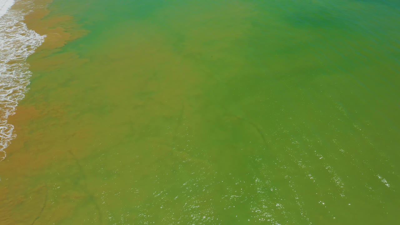 Aerial frame from San Jose Del Cabo, Mexico showing yellow-green water, orange-brown clouding near the left surf line, bright surface glints, and no visible people.