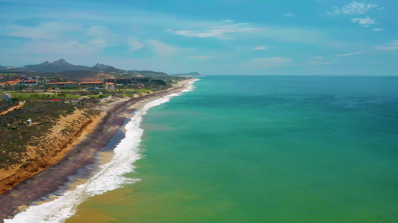 Aerial view in San Jose Del Cabo, Mexico with the surf line slightly farther down frame, green-blue ocean to the right, tan shore and inland buildings to the left, and no visible people.