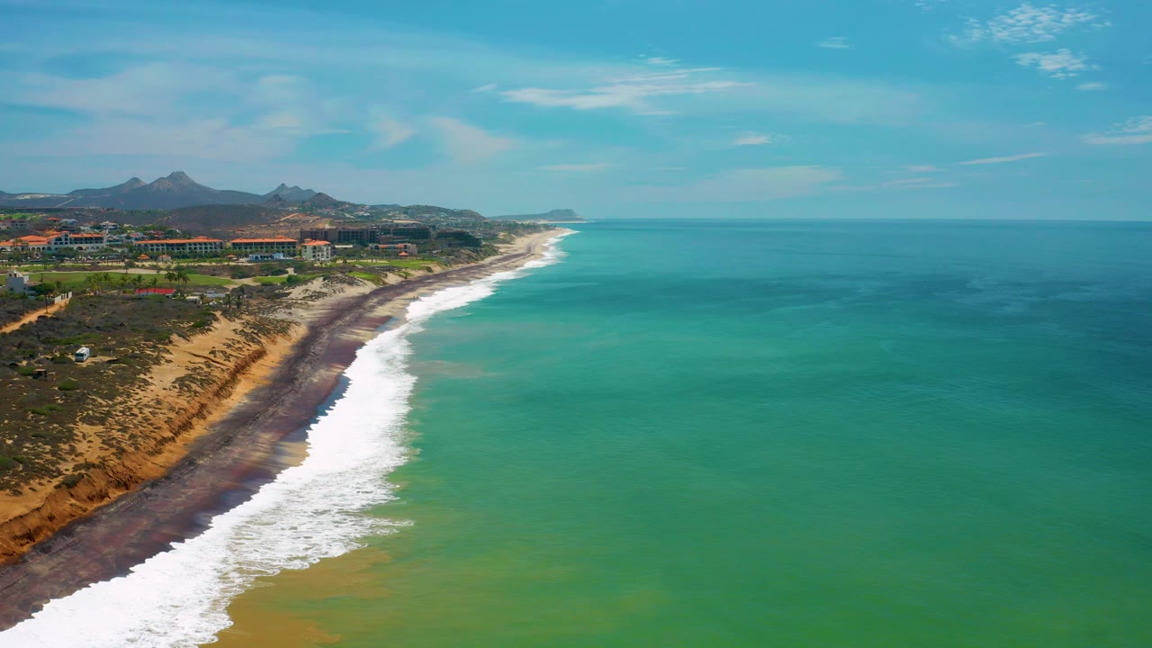 Aerial San Jose Del Cabo, Mexico coastline frame with foamy waves filling the lower left, green water across the center, a dark shore edge, distant buildings and hills, and no visible people.