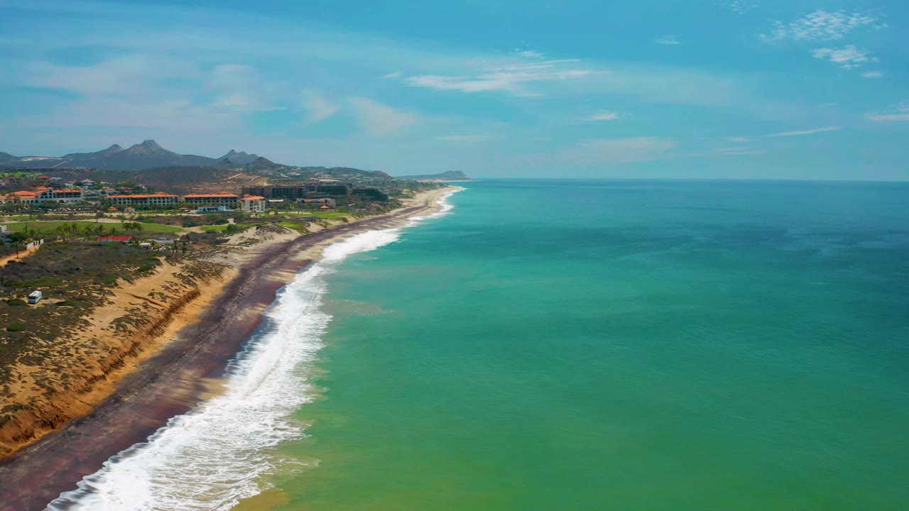 San Jose Del Cabo beach and ocean seen from a high offshore angle, with foamy waves tracing the shore, low buildings inland, mountains beyond, and no visible people.