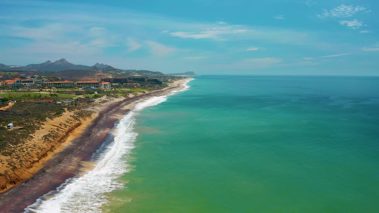 A wide San Jose Del Cabo coastal frame with more open turquoise water filling the right side, the surf line running toward the horizon, inland buildings, mountains, and no visible people.