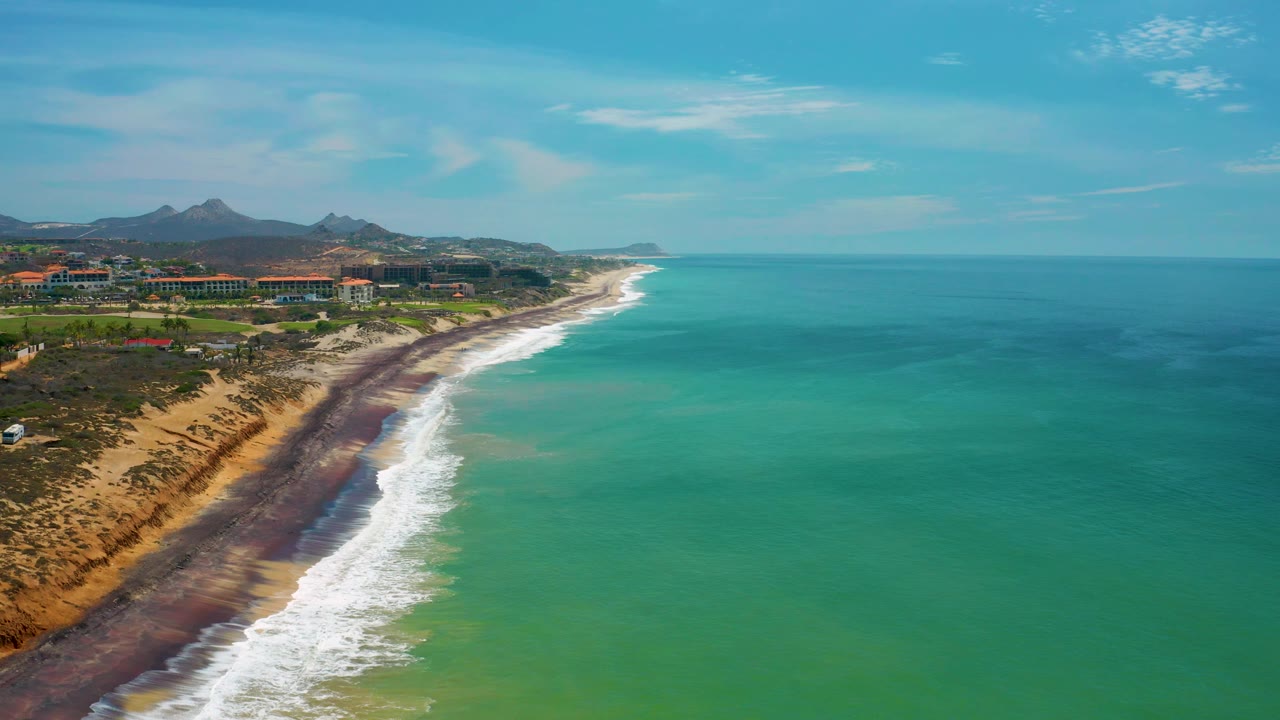 San Jose Del Cabo coastline from above with the beach crossing the lower frame, white waves pushing onto a dark shoreline band, resort grounds inland, mountains behind, and no visible people.