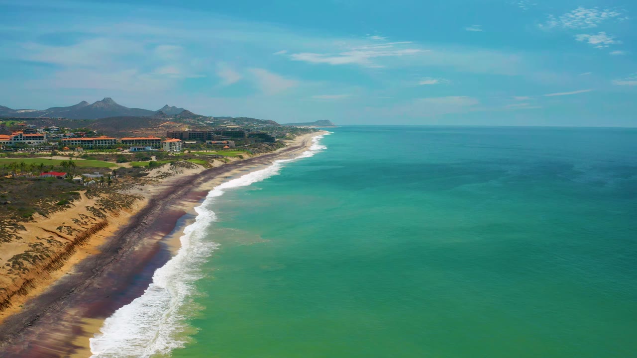 Elevated San Jose Del Cabo beach view with the white surf band slightly farther down the diagonal shoreline, green-blue water filling the foreground and right side, and no visible people.
