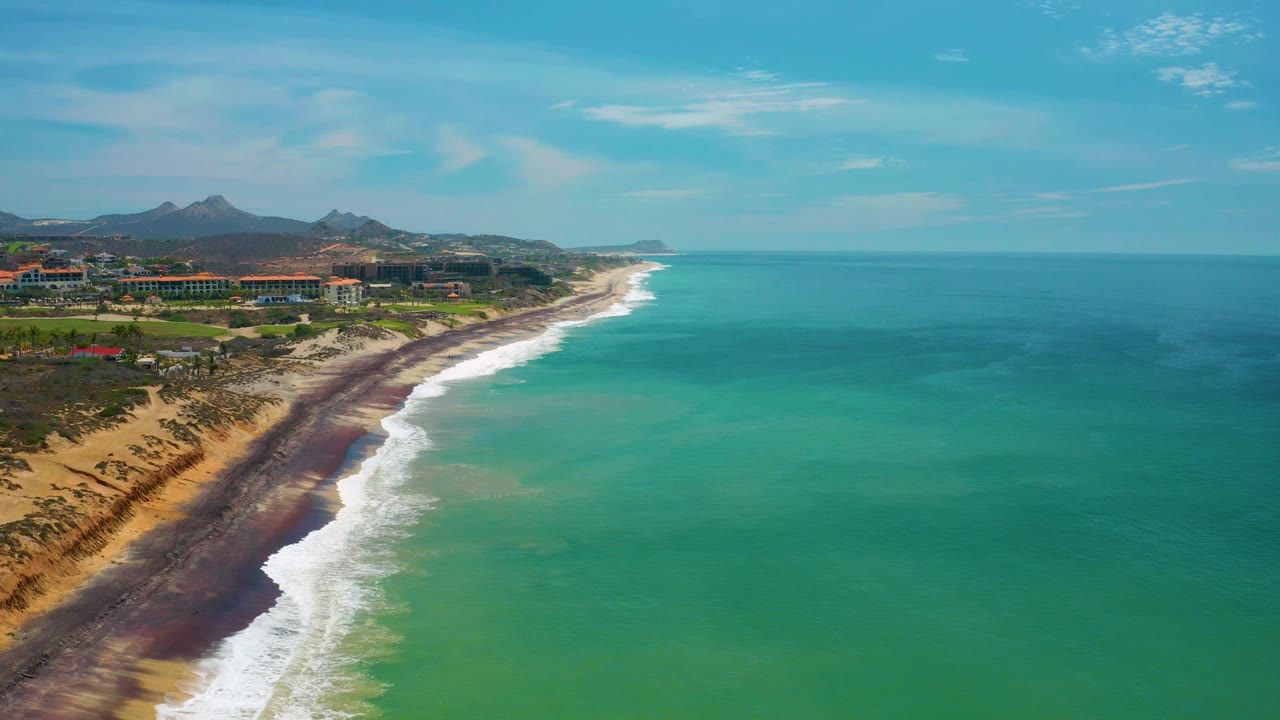 Aerial view of the San Jose Del Cabo shore with a long white surf edge, dark shoreline strip, developed coast at left, distant hills, open blue sky, and no visible people.