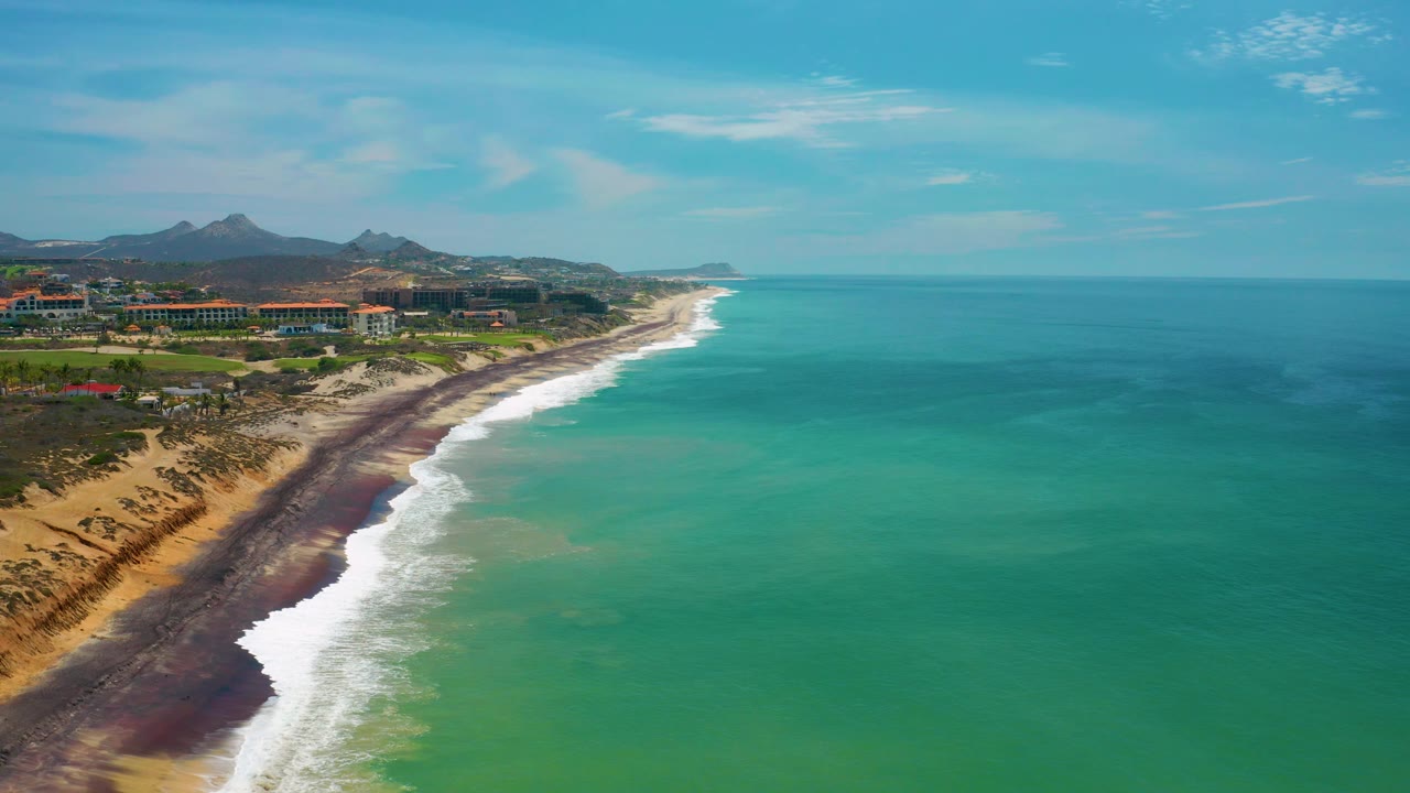 San Jose Del Cabo coastline from above, showing white waves along the left edge of the turquoise sea, a narrow dark shore band, sandy beach, low buildings, hills, and no visible people.