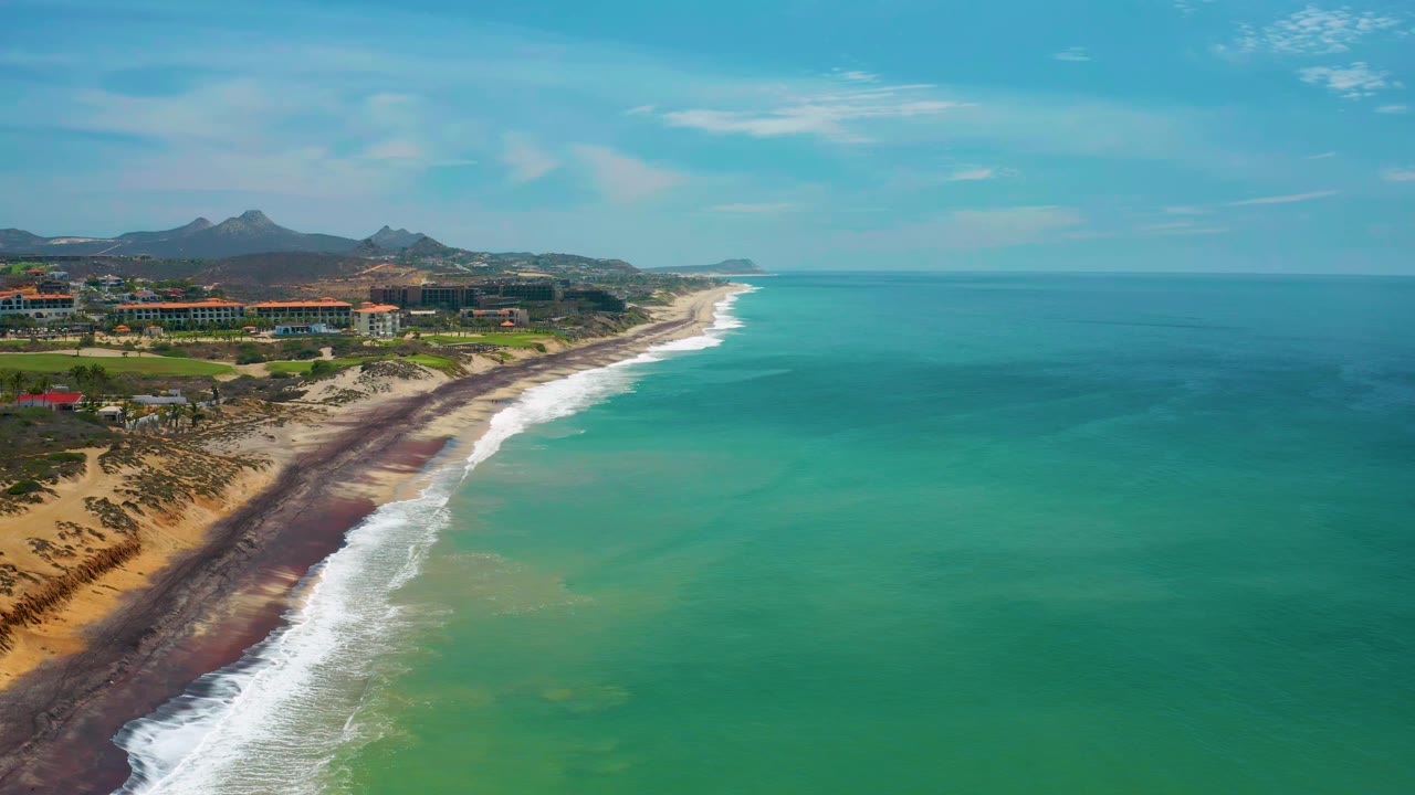 The second frame keeps the San Jose Del Cabo shoreline diagonal, with white waves breaking along the beach, a broad field of blue-green water, inland buildings, distant mountains, and no visible people.