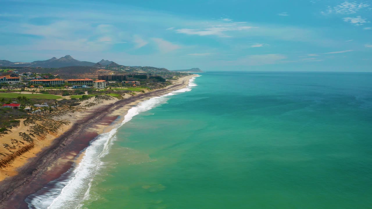 At 0:02 the aerial view shows more open ocean to the right, white surf curling against the San Jose Del Cabo beach, a strip of coastal development inland, mountains beyond, and no visible people.