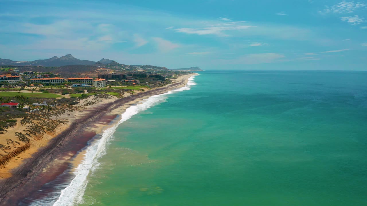 The 0:03 frame shows San Jose Del Cabo from above, with dunes and beach at lower left, white waves along the shore, turquoise water across the right side, inland buildings, and no visible people.