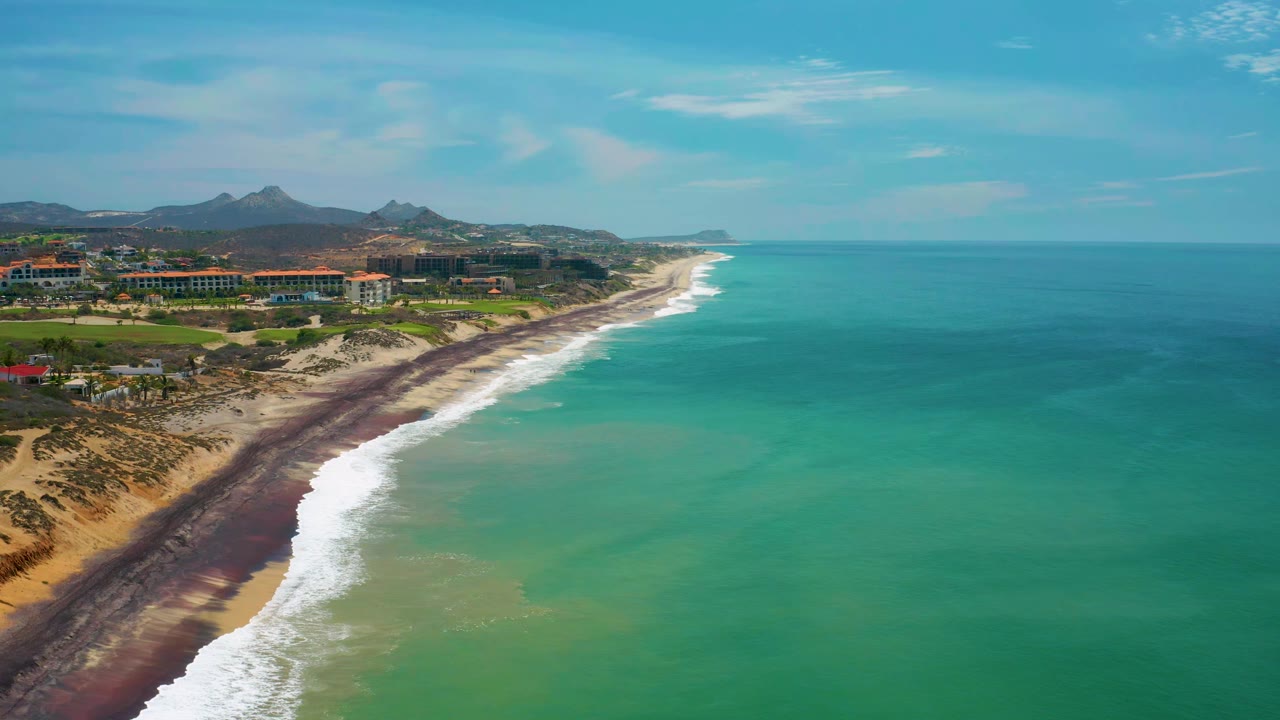 San Jose Del Cabo aerial coastline with surf curling onto the dark beach, turquoise and emerald water offshore, built-up shorefront, mountains, and no visible people.