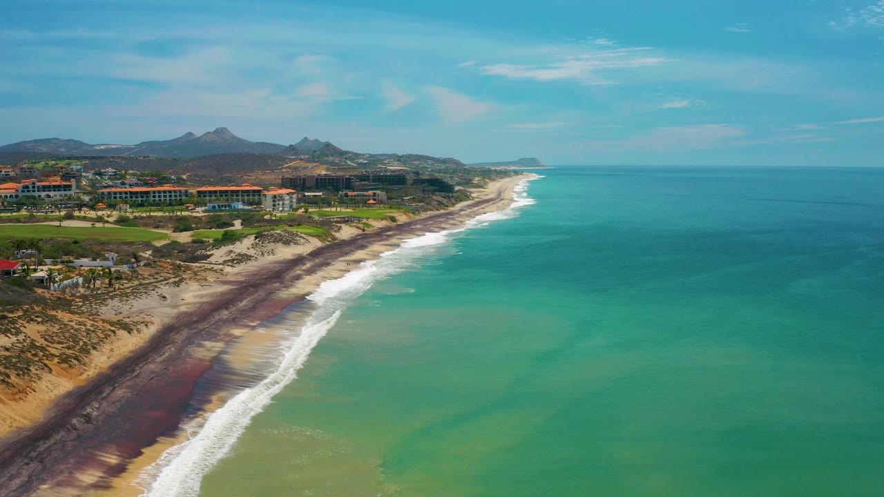 Aerial frame from San Jose del Cabo, Mexico showing turquoise water, a white surf line, sandy beach, low oceanfront buildings, distant hills, and no visible people.