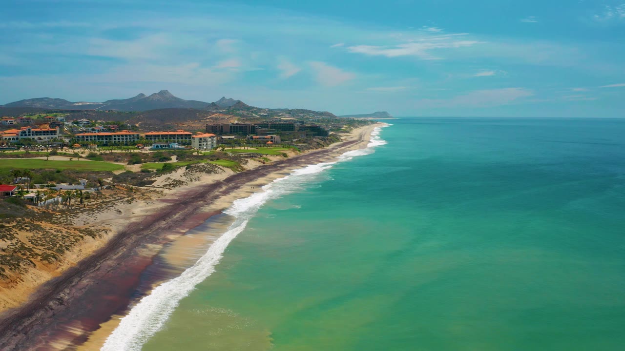 Aerial frame from San Jose del Cabo, Mexico showing more open turquoise water to the right, a long surf edge, beach and oceanfront buildings to the left, distant hills, and no visible people.