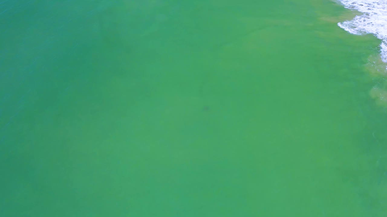 Drone-style view over San Jose Del Cabo sea water with a faint dark spot below the surface and small surf texture at the upper right, with no visible people.