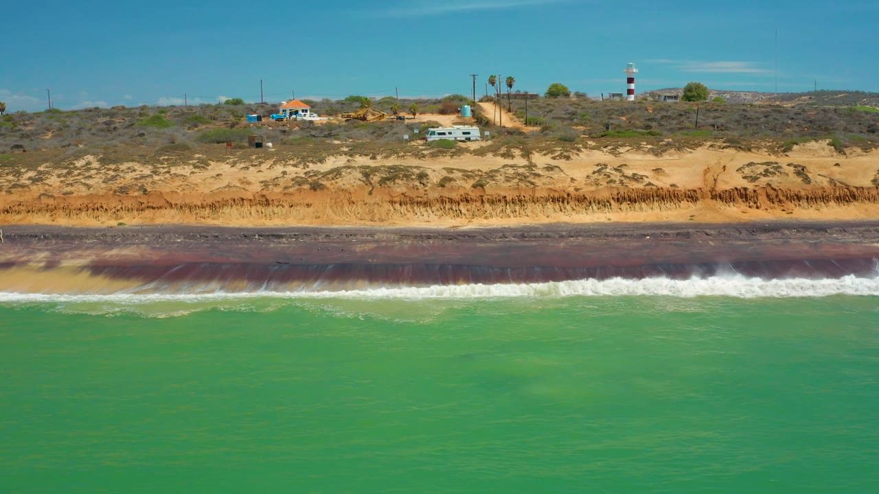 Aerial shoreline view in San Jose Del Cabo, Mexico, with broad turquoise water, a narrow surf break, dry sandy bluff, sparse vegetation, a striped tower, and no visible people.
